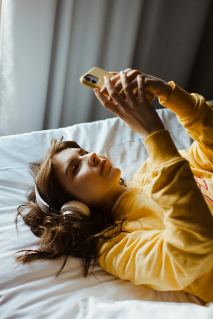 Young woman in a yellow sweater enjoying music on her phone while lying in bed.